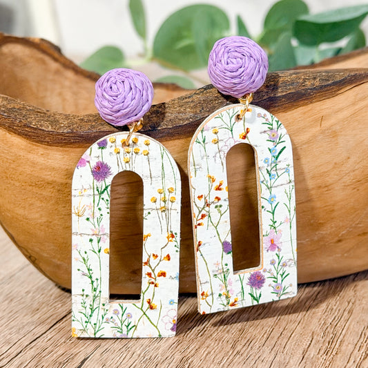 Lightweight leather arch earrings with a distressed white wildflower print and lavender raffia toppers displayed on a wooden bowl.