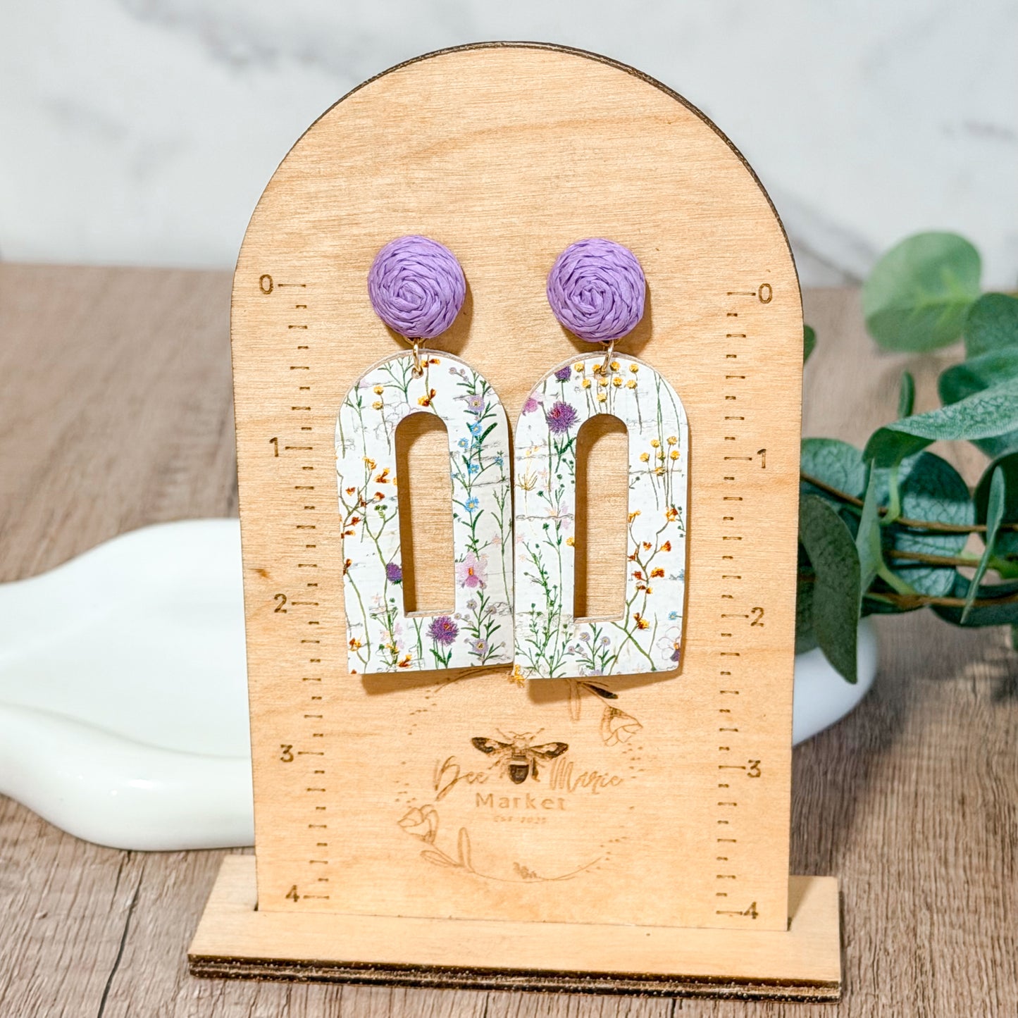 Lightweight leather arch earrings with a distressed white wildflower print and lavender raffia toppers displayed on a wooden stand.