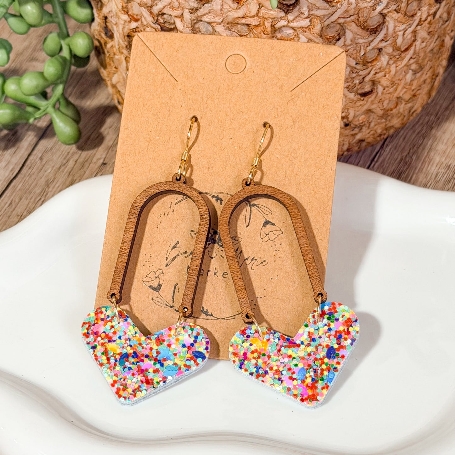 Colorful heart-shaped earrings on a brown card with a wooden surface and plant in the background