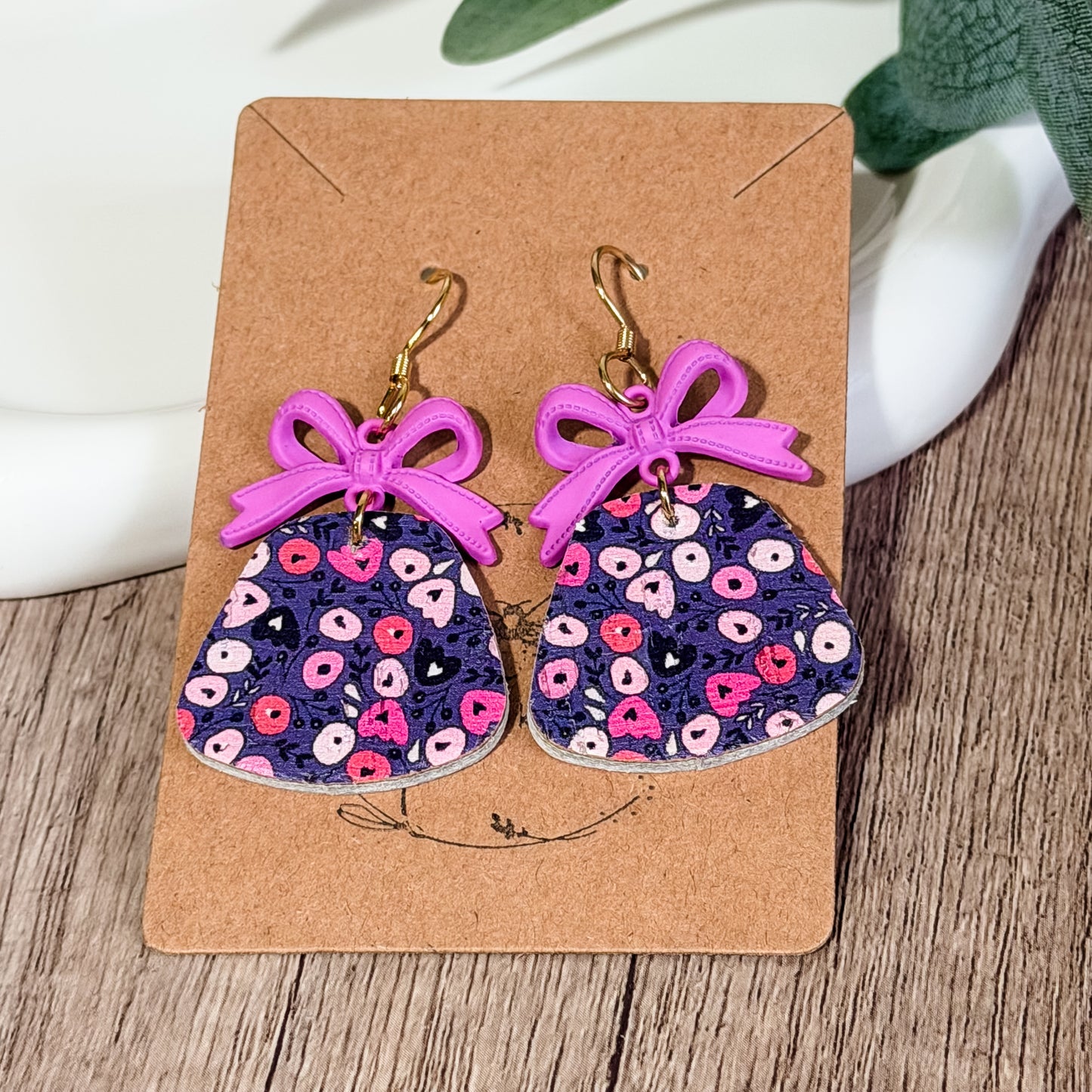 Purple floral leather earrings with lavender bow charms displayed on a kraft earring card on a wood background.