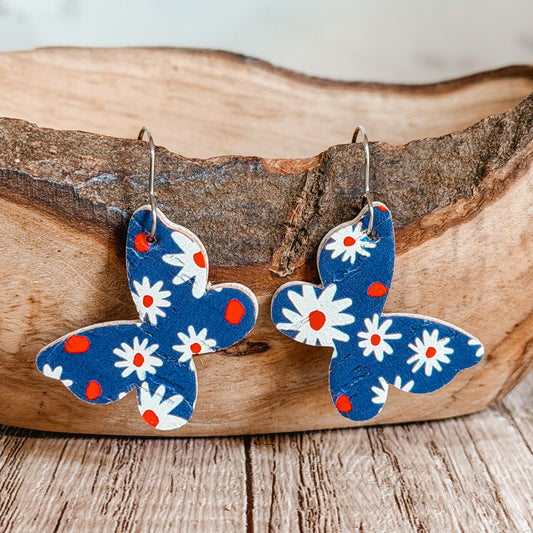 Handmade blue butterfly earrings with white daisy floral print and red accents displayed on a rustic wood bowl. Lightweight patriotic summer earrings with stainless steel hooks.