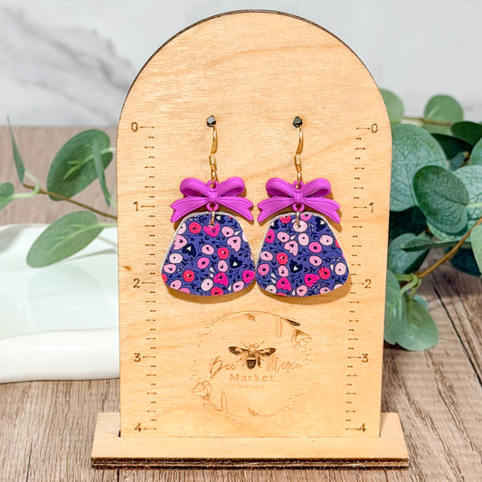 Purple floral leather earrings with lavender bow charms hanging on a wooden earring display stand engraved with the Bee Marie Market logo.