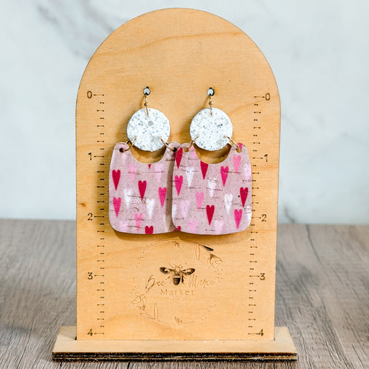 Pair of earrings with heart design on a wooden stand with measurement scale.