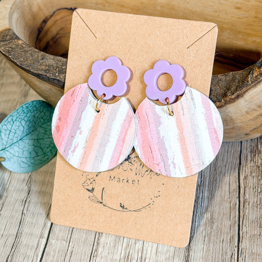 Pair of earrings with purple flower tops and pink striped bottoms on a brown card, placed on a wooden surface.