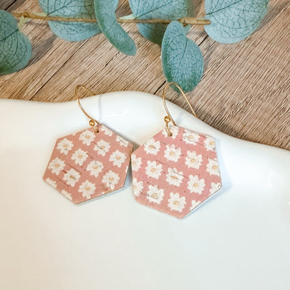 Hexagonal earrings with floral pattern on a white surface with green leaves in the background