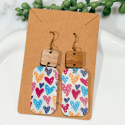 Colorful heart-patterned earrings on a brown card with greenery in the background