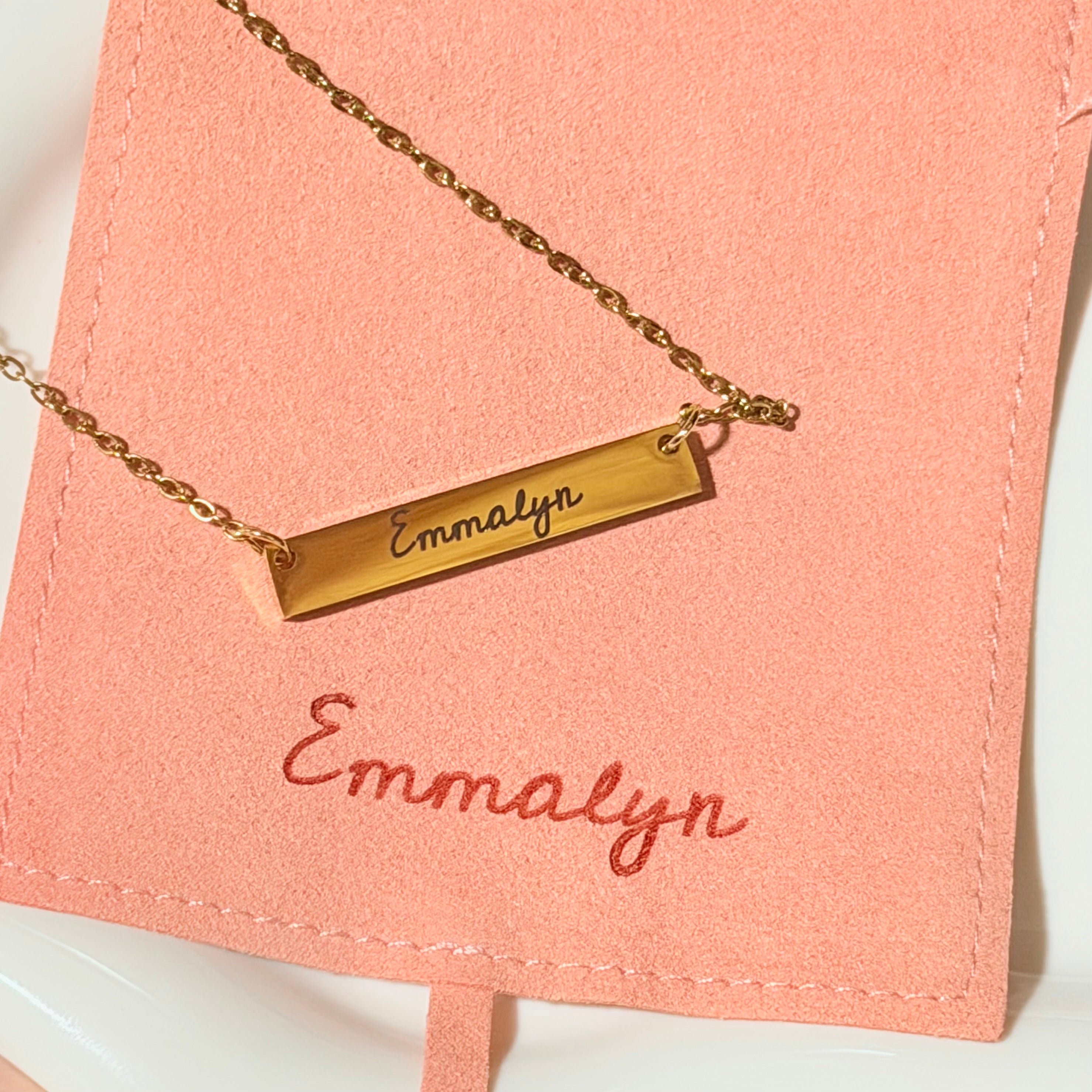 Gold necklace with a bar pendant on pink fabric with 'Emmalyn' engraved.