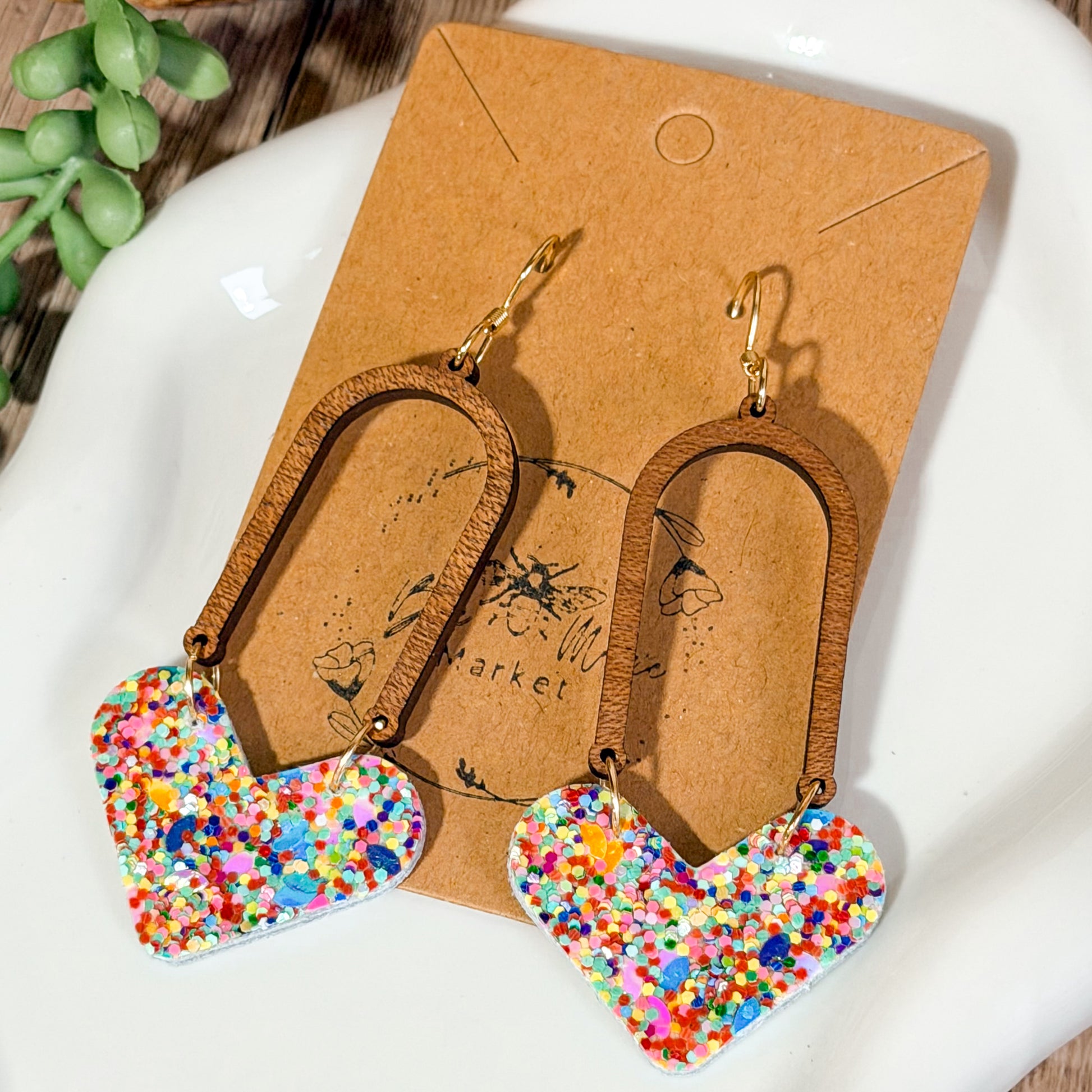 Alt text: Colorful confetti heart earrings hanging from wooden U shaped arches on a wooden earring display.