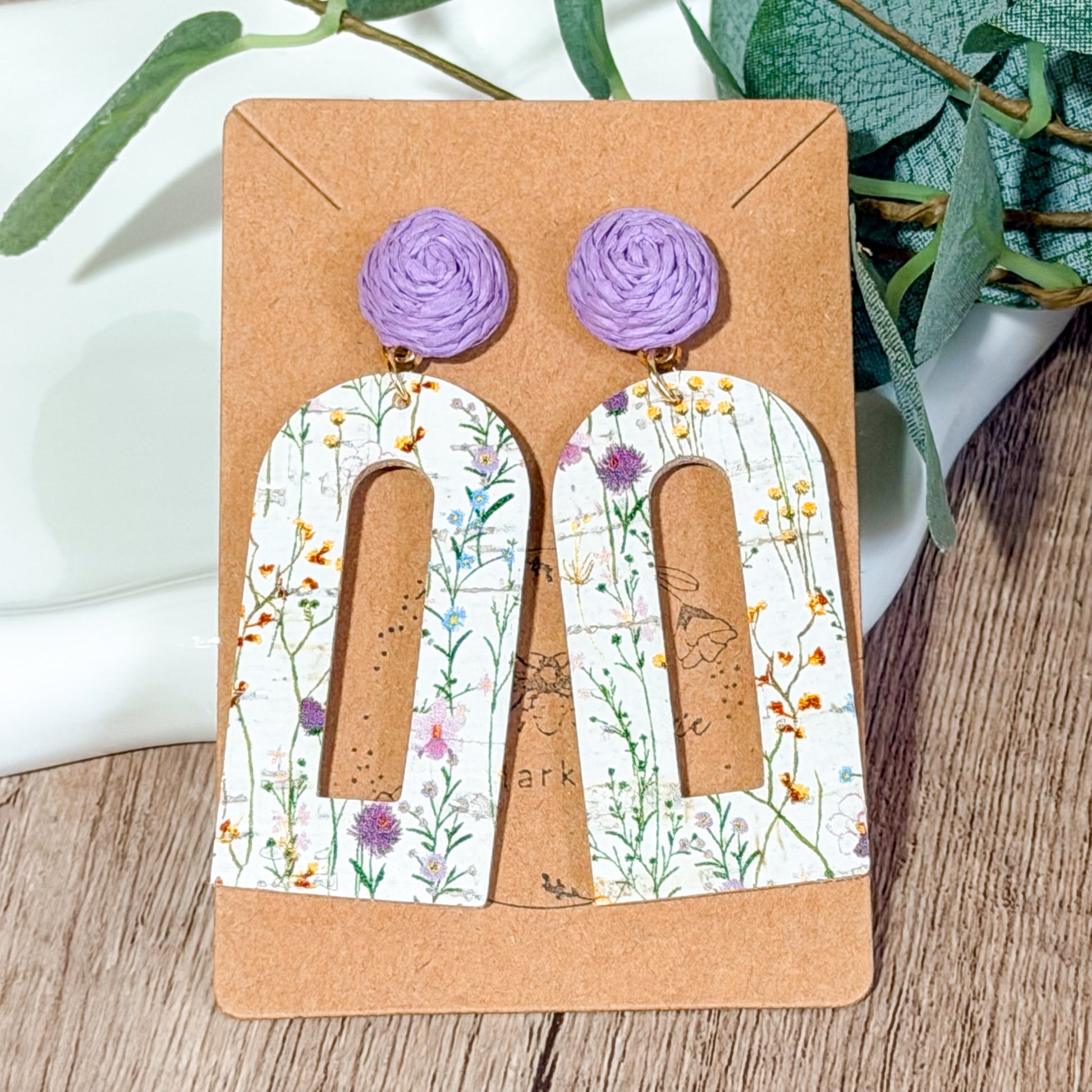 Lightweight leather arch earrings with a distressed white wildflower print and lavender raffia toppers displayed on a wooden card.