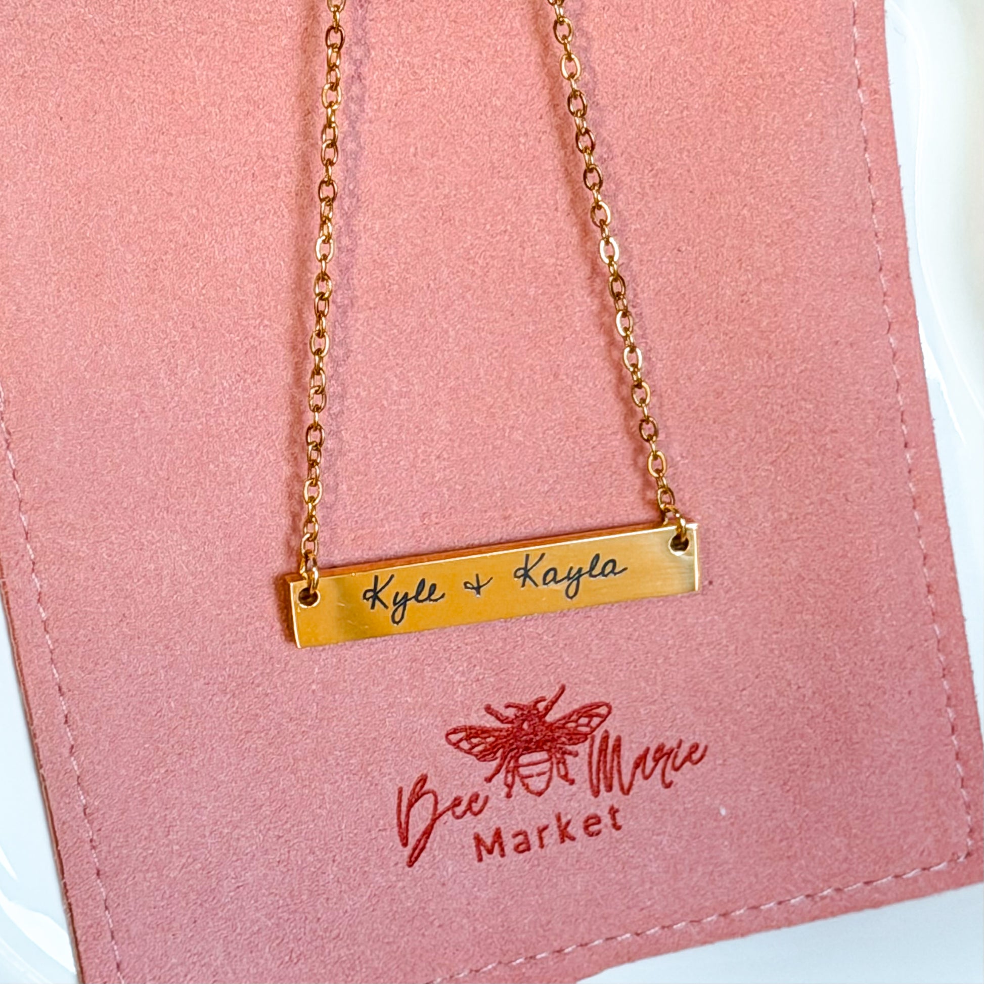 Gold necklace with engraved names on a pink card with 'Bee Marie Market' logo
