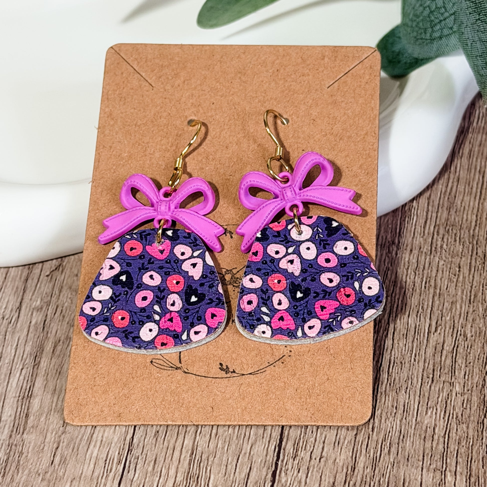 Purple floral leather earrings with lavender bow charms displayed on a kraft earring card on a wood background.