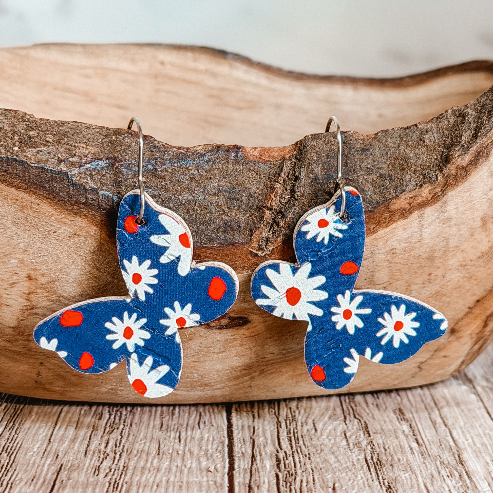 Handmade blue butterfly earrings with white daisy floral print and red accents displayed on a rustic wood bowl. Lightweight patriotic summer earrings with stainless steel hooks.