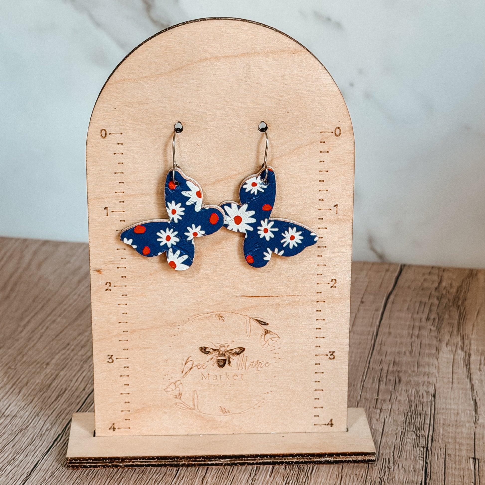 Handmade blue butterfly earrings with white daisy floral print and red accents displayed on a wood display. Lightweight patriotic summer earrings with stainless steel hooks.