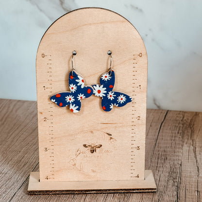 Handmade blue butterfly earrings with white daisy floral print and red accents displayed on a wood display. Lightweight patriotic summer earrings with stainless steel hooks.