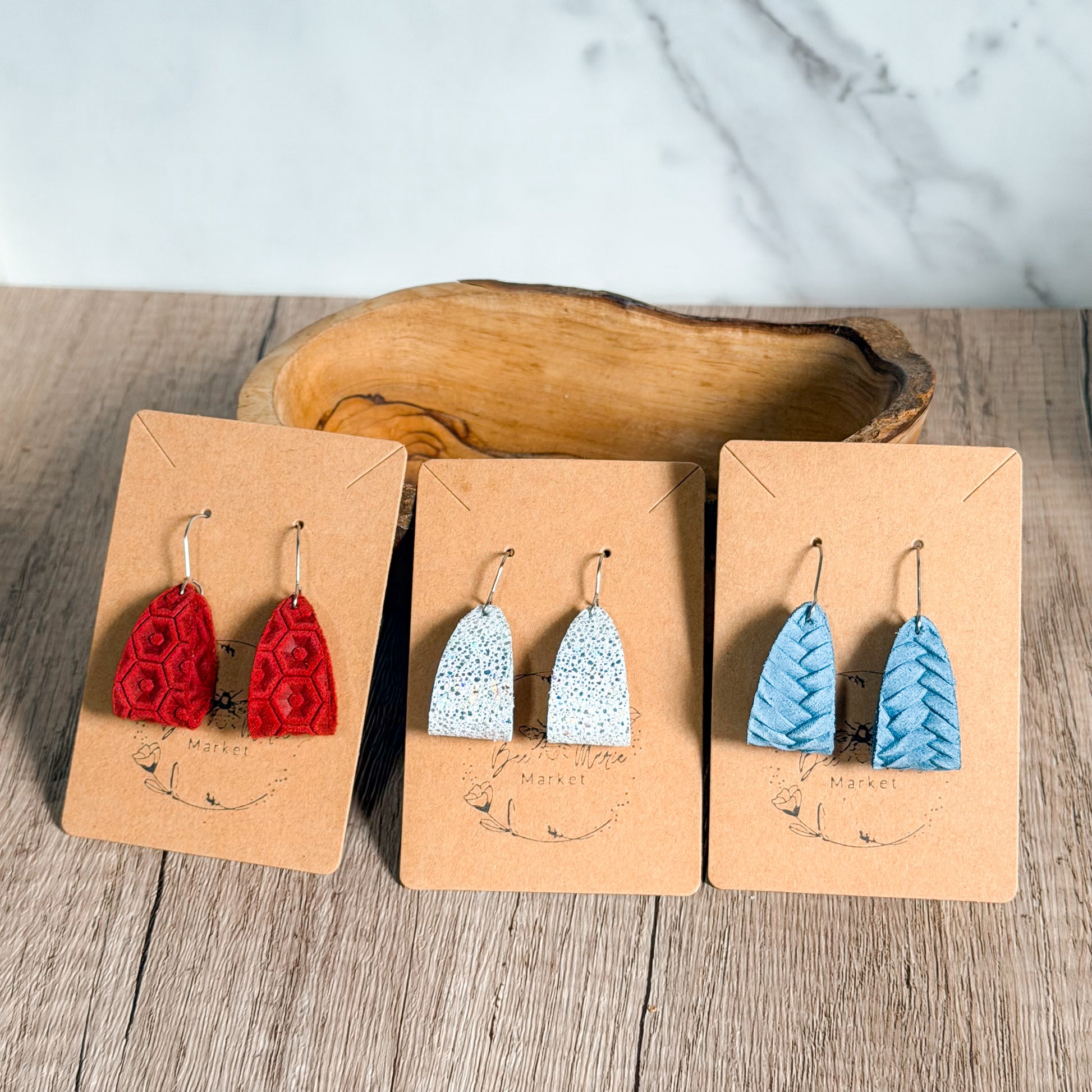 Three pairs of earrings on brown display cards with a wooden bowl in the background.