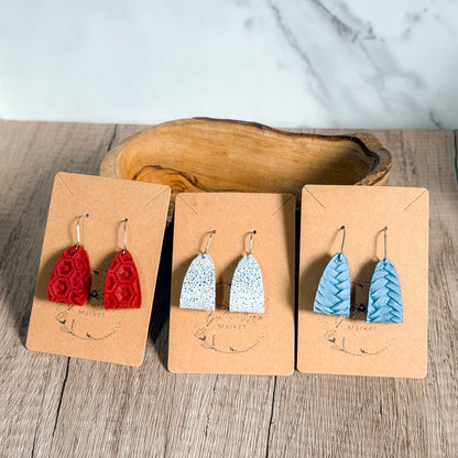 Three pairs of earrings on brown display cards with a wooden bowl in the background.