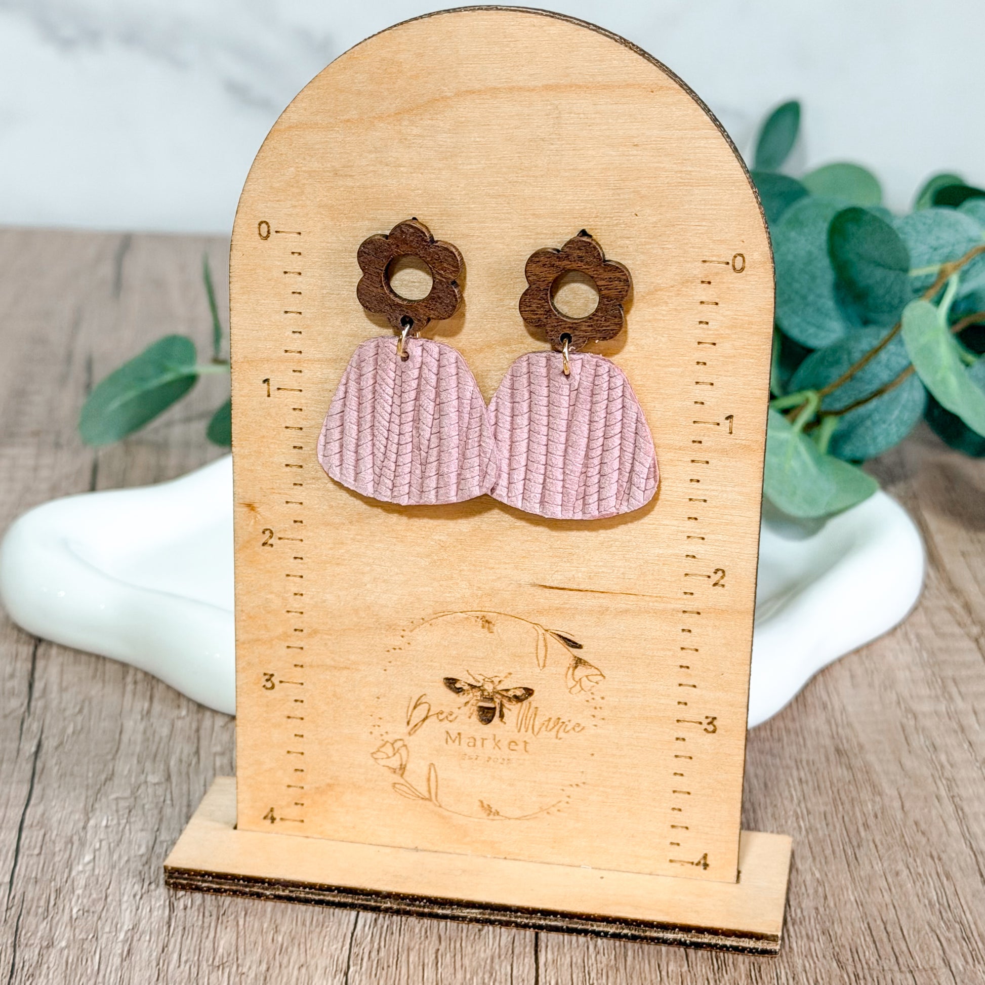 Light pink textured leather earrings with wooden flower studs displayed on a wooden Bee Marie Market stand.