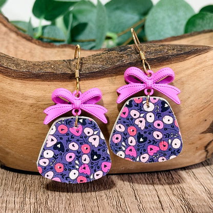 Purple floral leather earrings with lavender bow charms styled inside a wooden bowl with greenery in the background.