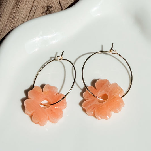 Peach-colored flower-shaped earrings on gold hoop earrings on a white surface.