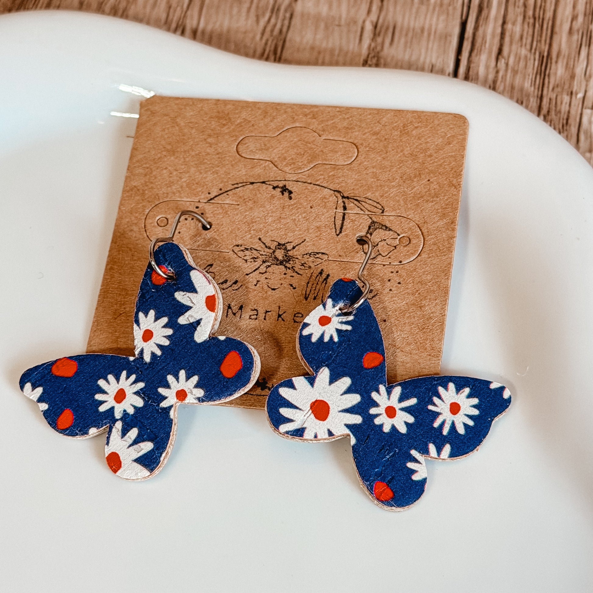 Handmade blue butterfly earrings with white daisy floral print and red accents displayed on a craft jewelry card. Lightweight patriotic summer earrings with stainless steel hooks.