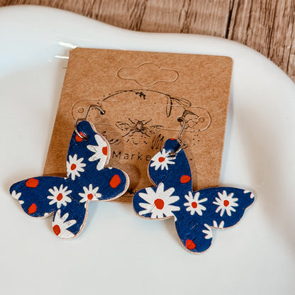 Handmade blue butterfly earrings with white daisy floral print and red accents displayed on a craft jewelry card. Lightweight patriotic summer earrings with stainless steel hooks.
