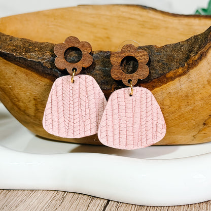 Light pink textured leather earrings with wooden flower studs displayed on a wooden bowl.