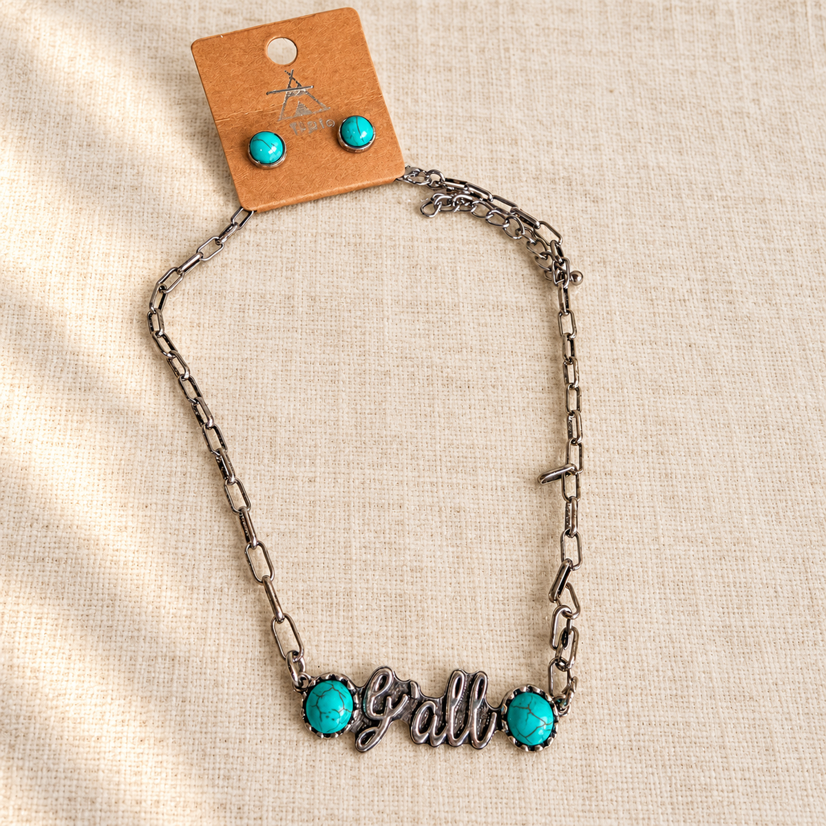 Full silver y'all necklace with turquoise accents and matching turquoise stud earrings on cream linen