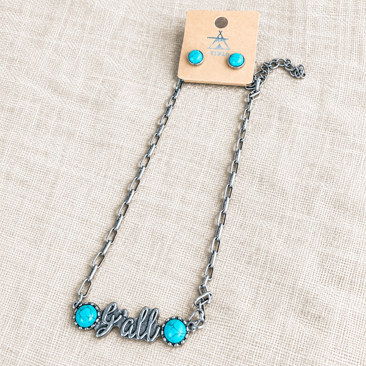 Silver y'all necklace and turquoise stud earrings on cream linen