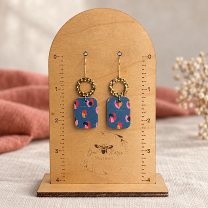 Teal abstract floral leather earrings with hammered gold hoops on a measuring stand